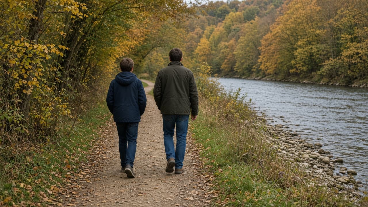 A youth and an adult walk a river-valley path, seen from behind, in daylight among autumn trees. They wear ordinary jackets.